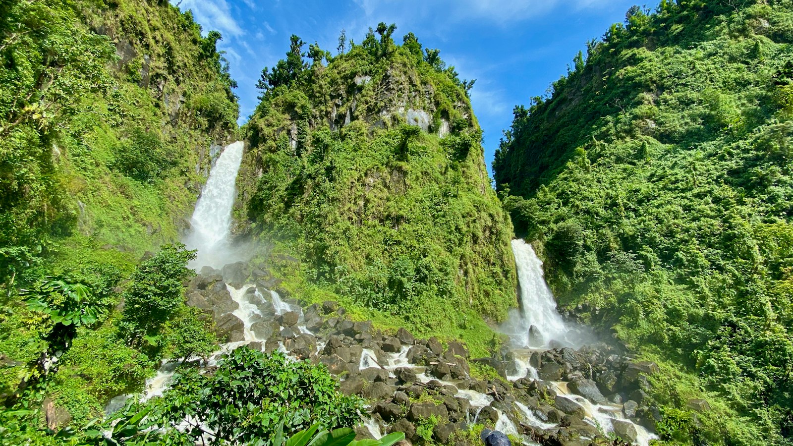 Twin waterfalls dropping through green mountainside forest in Dominica under a blue sky