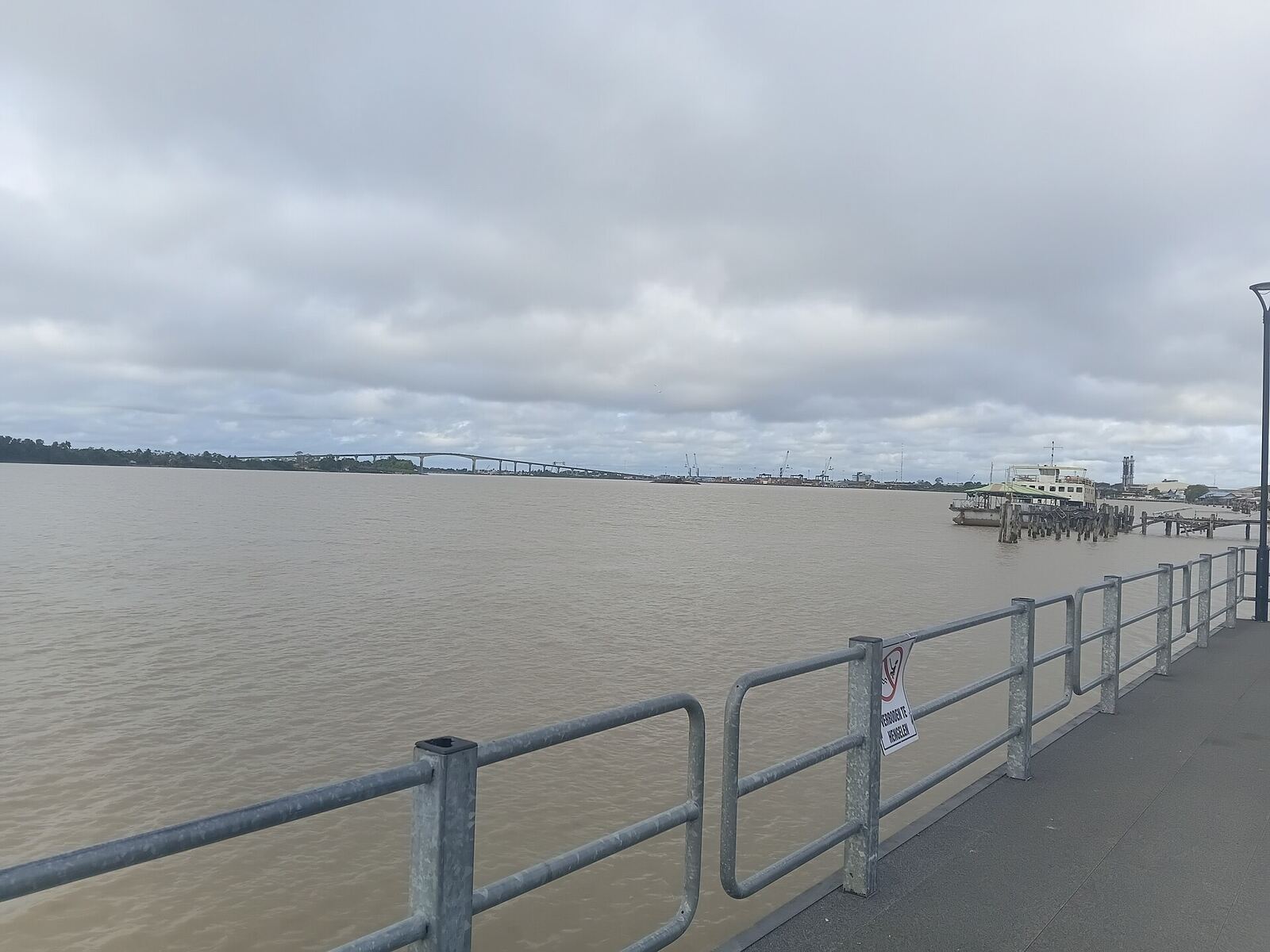 Brown water of the Suriname River beside the Waterfront promenade in Paramaribo under an overcast sky, a bridge far off